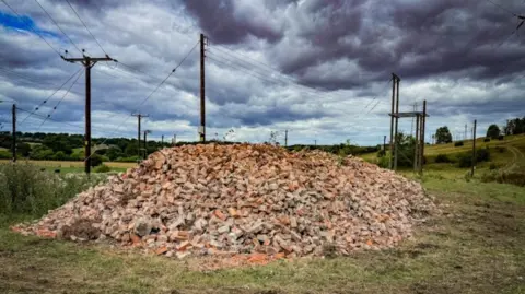 A massive pile of bricks in a field with telegraph poles behind. There are clouds in the blue sky.