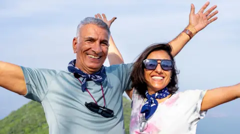 Anita Rani and her dad Bal standing together on a mountain, smiling and with their arms outstretched