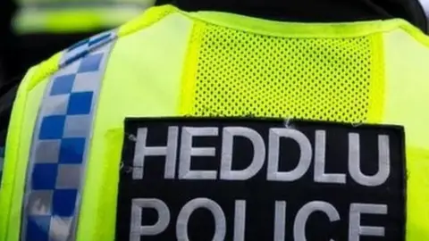 The badge of Police in Wales. White text on black badge on a yellow background. The text says Heddlu (in Welsh) which translates to Police, which is underneath.