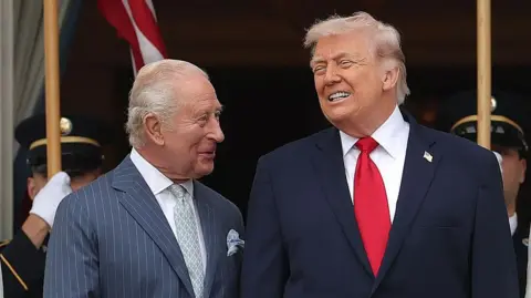 King Charles looking at Donald Trump, both wearing suits and smiling.