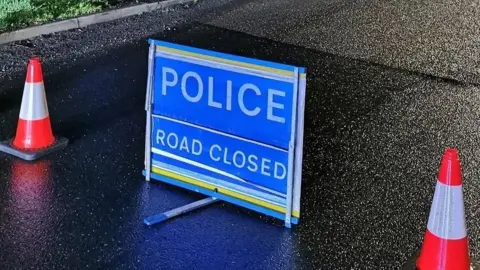 A road closed sign. The sign is blue and placed on a black tarmac road. There is an orange and white cone either side of the sign. 