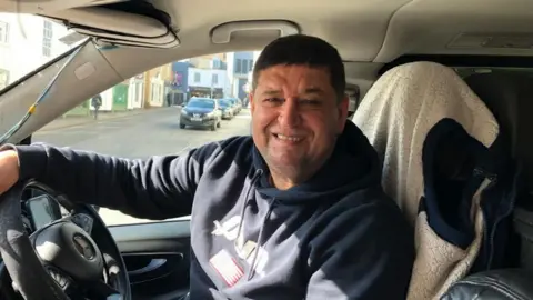 Vikki Irwin/BBC Haydar Bayir is sitting in his taxi and is smiling at the camera. He has dark brown hair and is wearing a hooded jumper which is blue.