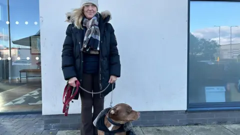 A woman in a light grey woollen hat, chequered scarf and black cold stands by a wall holding a brown bulldog on a lead. 