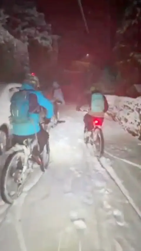 Cyclists bike down a narrow snow covered street in whilst snow falls