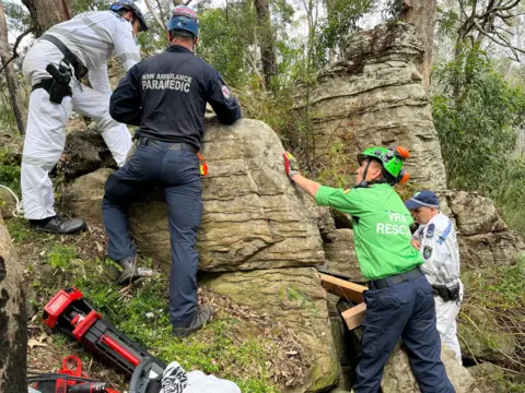 NSW Ambulance Vier mannen - een politieagent, een paramedicus, een man in een wit pak en een andere in een groene top met de tekst VAL Rescue - staan ​​naast een groot rotsblok in een bos. Er zijn stukken hout te zien die gebruikt zijn om de rotsen veilig te stellen, evenals mechanische uitrusting