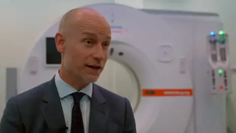 A slim, bald man in suit stands in front of a scanning machine in a hospital.