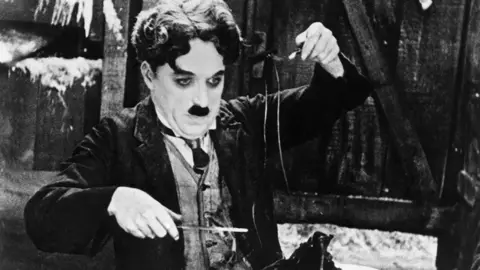 Getty Images A black and white still of Charlie Chaplin from The Gold Rush. He is wearing a dark suit and time with a white shirt. He is lifting a shoelace with a fork, with a knife in his right hand