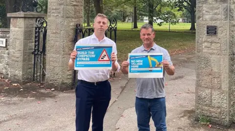 Nick Evans Tidenham Councillor Nick Evans wearing dark trousers and a light shirt, holding up a sign that says 'build the Chepstow bypass'. He is standing beside another man beneath a large stone archway at the entrance to a park. 