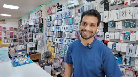 Muhammad Usman a man in a blue t-shirt, sat behind a desk and surrounded by phone accessories. He is smiling at the camera.