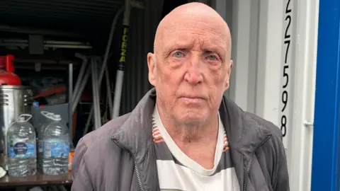 Gavin Kermack / BBC A bald man wearing a striped t-shirt under a grey jacket. He is standing in front of a container like the one in the above picture. It is open and bottles of water and miscellaneous equipment are inside.