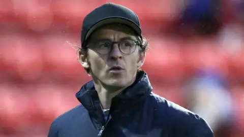PA Media A close up of Joey Barton. He is standing in a football stadium, and wearing a black jacket and cap, and glasses.