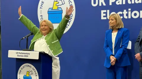 Pat Cullen stands  in front of a lectern with her hands raised above her head. She is smiling while to her right is Diana Armstrong, the UUP candidate in the election