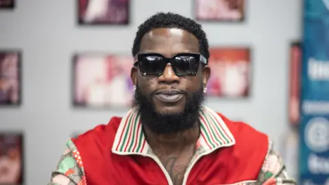Gucci Mane looks into the camera wearing sunglasses and a red shirt.