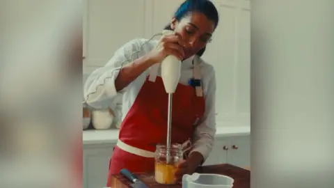 A woman with long, dark hair wearing a red apron over a white shirt blends liquid in a glass jar