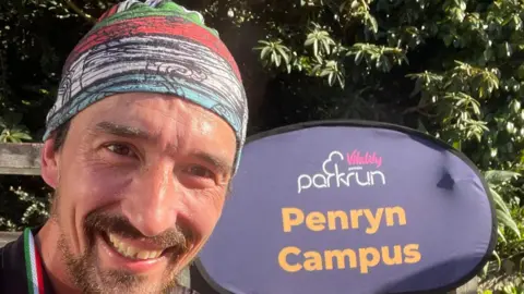 Adam 'Tango' Holland Adam Holland smiling and wearing a stripy bandana, stood in front of a purple Penryn Campus Parkrun sign.