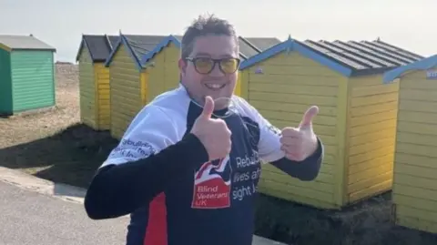 Wayne Pope wearing yellow sunglasses and a blue and white running shirt with Blind Veterans UK charity branding on it. He is running past a row of colourful beach huts and smiling at the camera, giving a double thumbs up.