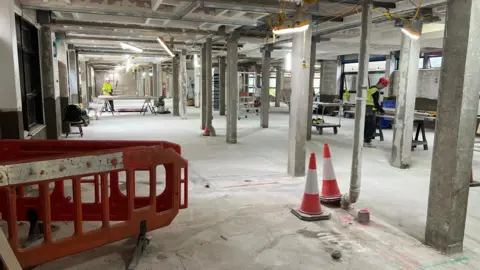 The image shows a building site in a former hospital ward where all the ceilings, floors and walls have been stripped out. There are workmen in hardhats working to deal with the problems with crumbly concrete.