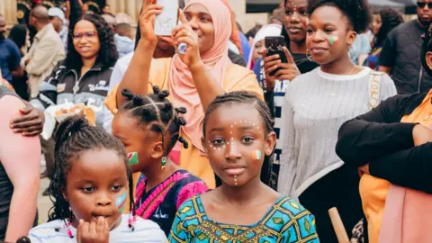 A group of women and children at a festival. Three children can be seen at the front of the crowd, with their faces painted.
