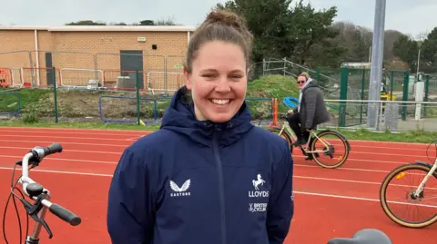 A young woman wearing a blue waterproof jacket, stood in front of some people on bikes and a bike on its stand.