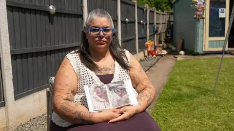 A woman with grey/dark hair and blue framed glasses. She sits in a garden and wears a purple dress and white crochet top.