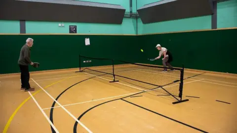 Cotswold District Council An older man and older woman play pickleball in a leisure centre. The woman is slightly bending down to hit the ball back to the man over the net. The walls are green and the floor is a light wood colour with black, yellow and white markings.