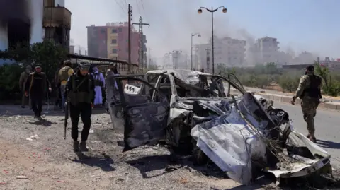 Bedouin fighters walk near a damaged car in Suweida, following the Syrian presidency's announcement of a ceasefire, 19 July 