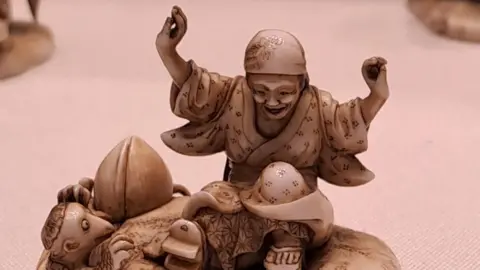 Bradford District Museums and Galleries Intricately carved ivory netsuke depicting a seated figure with raised arms, wearing a patterned robe and head covering. In front of the figure lies a large peach and a small creature resembling a kappa, with detailed scales and expressive features.