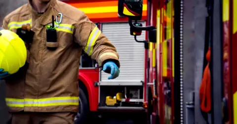 A generic image of a firefighter's body. He is in a brown uniform with yellow and white trim and is wearing blue gloves and carrying a yellow firefighter helmet. Behind him and to the side are fire engines. 