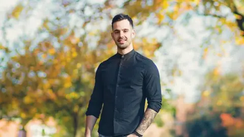 Bavington family Robert Bavington with short dark hair and stubble. Stood in front of trees with yellowing leaves in autumn. He's wearing jeans and a navy shirt.