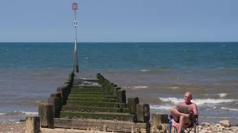 A man in black shorts sits with legs crossed reading a paper while sitting on a folding chair in front of the sea.