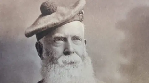 The black and white portrait of Charles Fraser-Mackintosh shows a elderly man in a bonnet with a bushy white beard. He has piercing eyes which are looking slightly away from the camera.