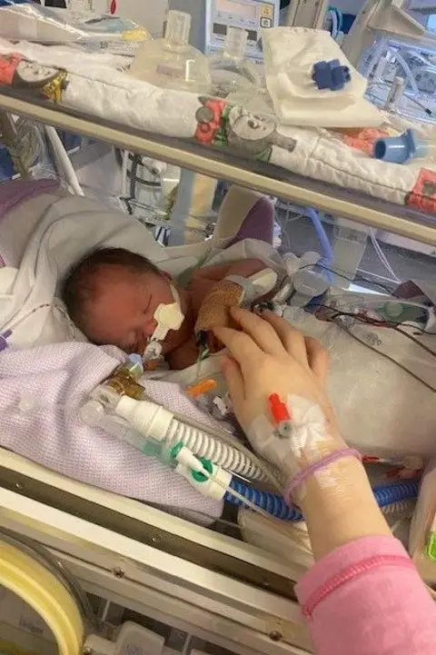 Family photo a woman's arm in a pink sleeve stretches out towards a newborn baby on a special care baby unit 