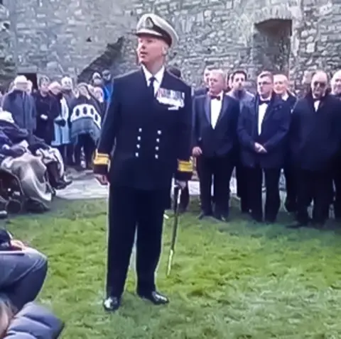 A man in a navy officer's white hat and blue blazer stands addressing a crowd against a castle wall. In his left hand he holds a ceremonial sword.