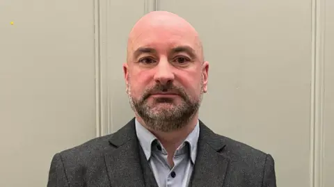 PCC Marc Jones he has a shaved head and dark beard he is wearing a charcoal three-piece suit with light grey shirt