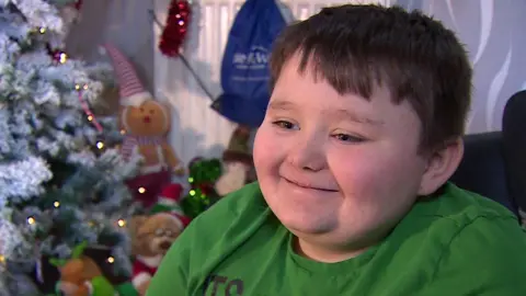 Jaxen is wearing a green Christmas T-shirt. He is pictured in front of his tree which has lights and soft toys in front of it. 