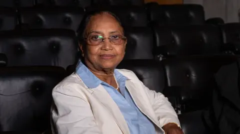 Author Banu Mushtaq pictured wearing a white overcoat and a powder blue shirt, smiling for the cameras. 