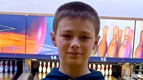 FAMILY Leo looking straight at the camera. Ten-pin bowling is behind him with an alley leading to a set of pins.
