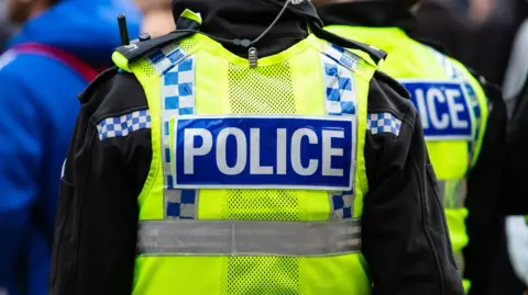 Getty Images Two police officers seen from behind wearing high-visibility yellow jackets with blue and white chequered markings and 'POLICE' patches on the back. The officer in the foreground has a radio clipped to the shoulder of a black fleece underneath, with a coiled earpiece wire visible. The image is taken in a crowd setting with other figures partially visible in the background.
