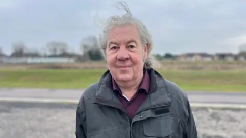 A man with long curly grey hair smiles at the camera. he is wearing a purple shirt and a blue coat and stands in front of a road and pavement. A strand of hair is blowing in the wind.