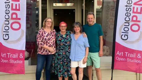 Deborah Harrison CIC directors of Gloucester Contemporary Artists, from left to right: Wendy Golding, Gilly Hill, Deborah Harrison and David Finch stand in front of a shop with 'Gloucestershire Open' banners on the side 