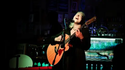 Gideon Lydiard Athena Aperta sings on stage, guitar in hand, the room behind her is dark and a spotlight illuminates her face and guitar. 