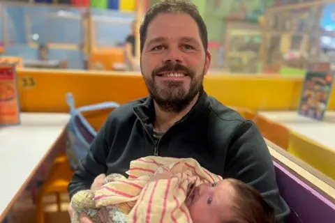 A man smiles as he holds a baby in his arms. He has short brown hair and a beard and wears a black fleece. The baby has brown hair and is wrapped in a yellow and pink striped shirt. They are in a seating area at a soft play centre. The background is out of focus.