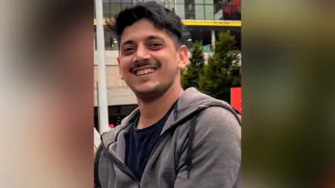 A man with short black hair and a short stubbly moustache and beard wears a grey zipped hoodie and black T-shirt while smiling for a photo.