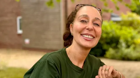 Cecile Roberts is sitting down on a bench outside. She is wearing a green T-shirt and is looking directly at the camera and smiling. Behind her is a brick building and some green foliage.