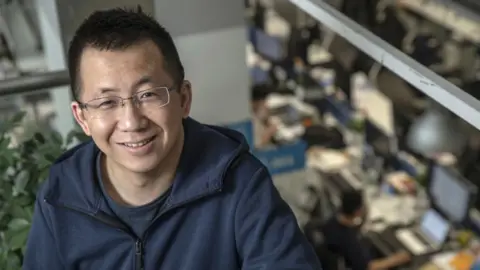 Getty Images Zhang Yiming, sitting in an office. Behind him you can see people working at their desks. He is smiling, wearing glasses and a laid-back look including a t-shirt and jacket.