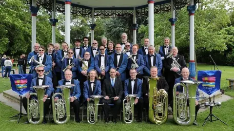 Southend-on-Sea City Council A brass band lined up in four rows in front of a bandstand. Members are holding their instruments and wearing matching uniforms of blue blazers with bow ties.