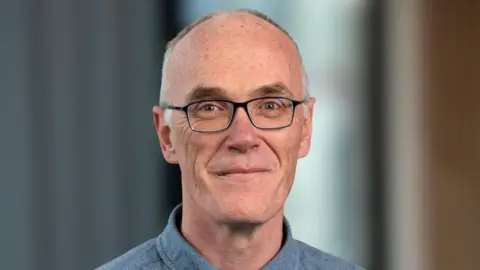 Ulster University Dr Eoin Magennis is smiling at the camera in a lecture hall wearing a pair of dark framed glasses. He has short grey/white hair and is wearing a blue/grey shirt. 
