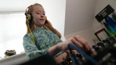 A young girl with rainbow headphones plays on DJ decks inside a house.