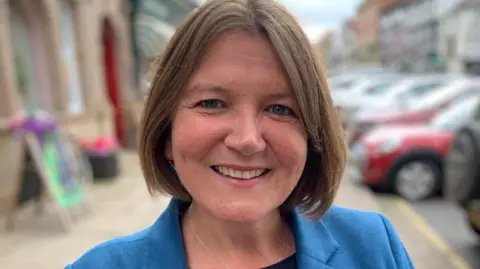 Ellie Chowns pictured in a blue jacket and black top. She has short brown hair and has been pictured on a pavement with cars behind her.