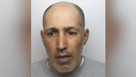 A mugshot of Rahman, who is unshaven, balding and wearing a grey jumper.